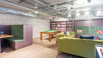 A modern lounge with a pool table, green cushioned seating, and a bookshelf.