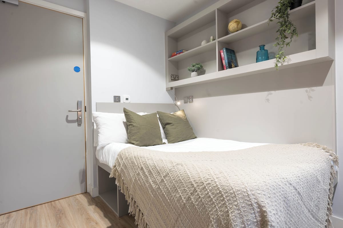 Un dormitorio con cama, mesita de noche, estante con libros y objetos decorativos, y una puerta.