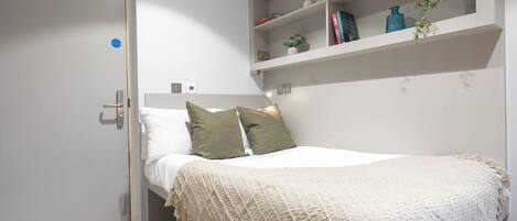 A bedroom with a bed, a bedside table, a shelf with books and decorative items, and a door.