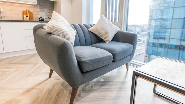 A modern living room with a grey sofa, two white cushions, a wooden coffee table, and a view of a cityscape through a large window. - City HideAway (Galway)