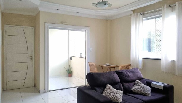 A modern living room with a dark sofa, a glass door leading to a balcony, and a dining area with a small table and chairs. - Apartment in Belém cop30/2025 (Coqueiro Ananindeua)