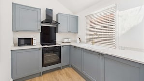 A compact kitchen with grey cabinets, a microwave, a toaster, a stove, and a window with blinds. - Host Stay Captain s View (Whitby)