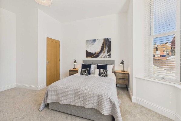 A bedroom with a bed, a wooden door, a painting on the wall, and a window with blinds. - Host Stay Holmwood (Whitby)