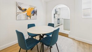 A dining area with a round table and four blue chairs, a modern abstract painting on the wall, and a view into the kitchen through an archway. - Host Stay Holmwood (Whitby)