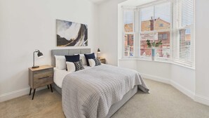 A bedroom with a bed, a nightstand, a lamp, a painting, and a window with blinds. - Host Stay Holmwood (Whitby)