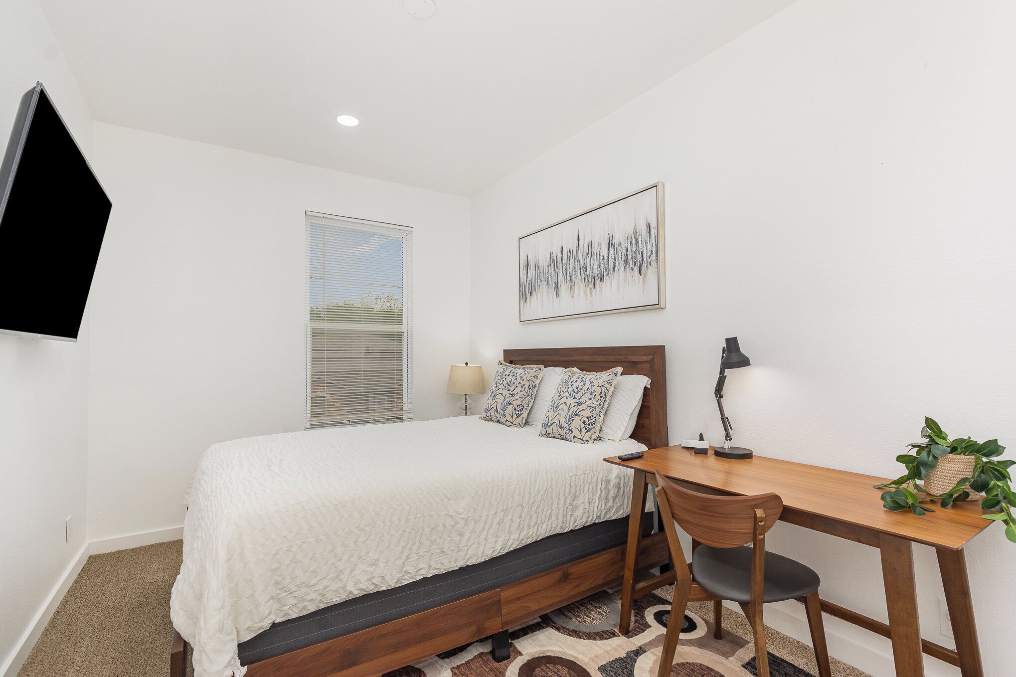 A bedroom with a bed, a desk, a chair, a lamp, a plant, and a painting on the wall.