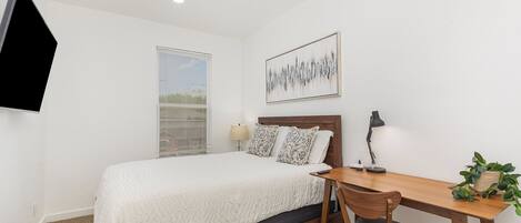 A bedroom with a bed, a desk, a chair, a lamp, a plant, and a painting on the wall.