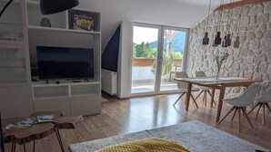 A modern living room with a flat-screen TV, a dining area with a table and chairs, and a large window offering a view of greenery. - Neckar View Apartment near Heidelberg | Balcony & Parking (Neckarsteinach)