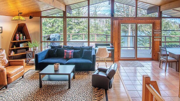 En rummelig opholdsstue med en stor sofa, et sofabord og en pejs. Der er en balkon med gelænder i træ og udsigt til det omkringliggende trælandskab.