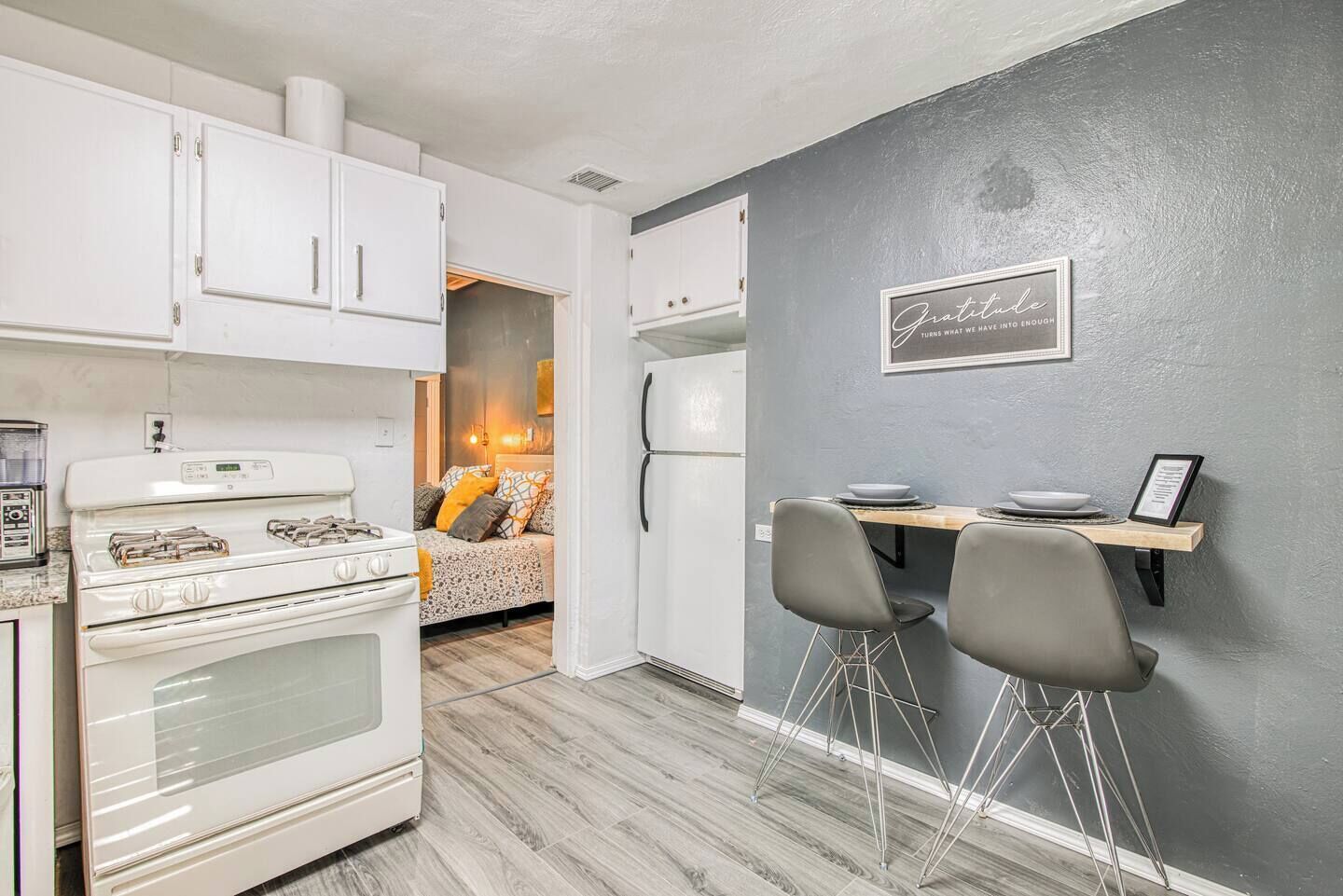 A compact kitchen with white cabinets, a stove, and a refrigerator. There's a dining area with two chairs and a small table. A framed quote is on the wall.