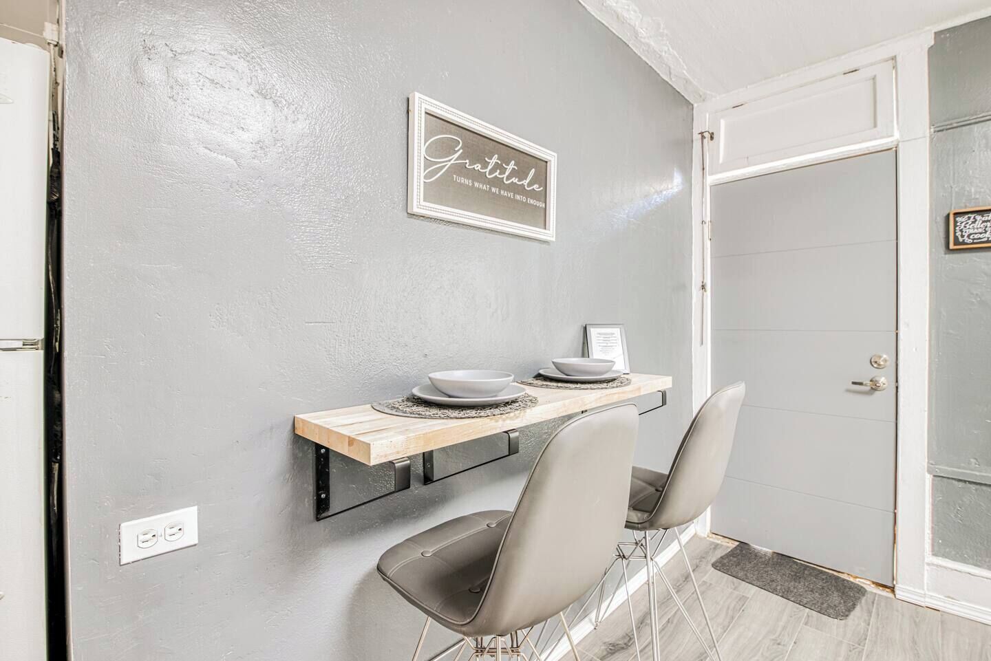 Un pequeño comedor con una pared gris, una repisa de madera con platos y dos sillas.
