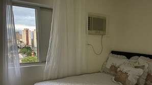 Un dormitorio con cama, cortinas blancas, ventana con vista a edificios y aire acondicionado instalado en la pared.