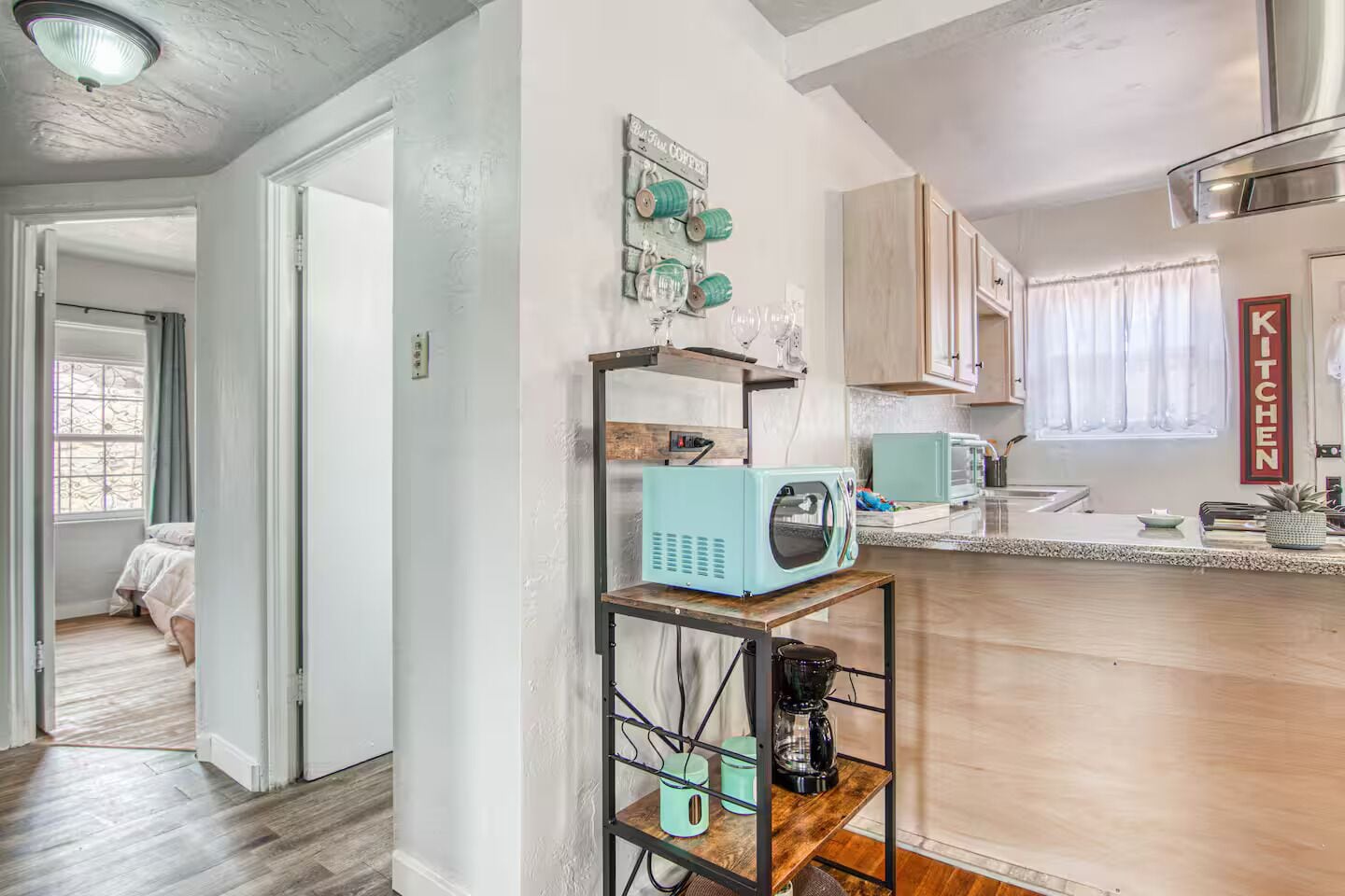 A kitchen with a teal microwave, a coffee maker, and a shelf with various items. There is a sign that says "Kitchen" on the wall.