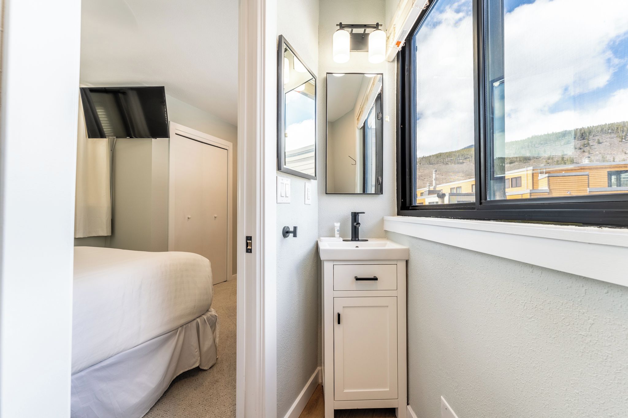 Ein Schlafzimmer mit einem Bett, einem weißen Schrank mit Spiegel und einem Fenster mit Blick auf Gebäude.