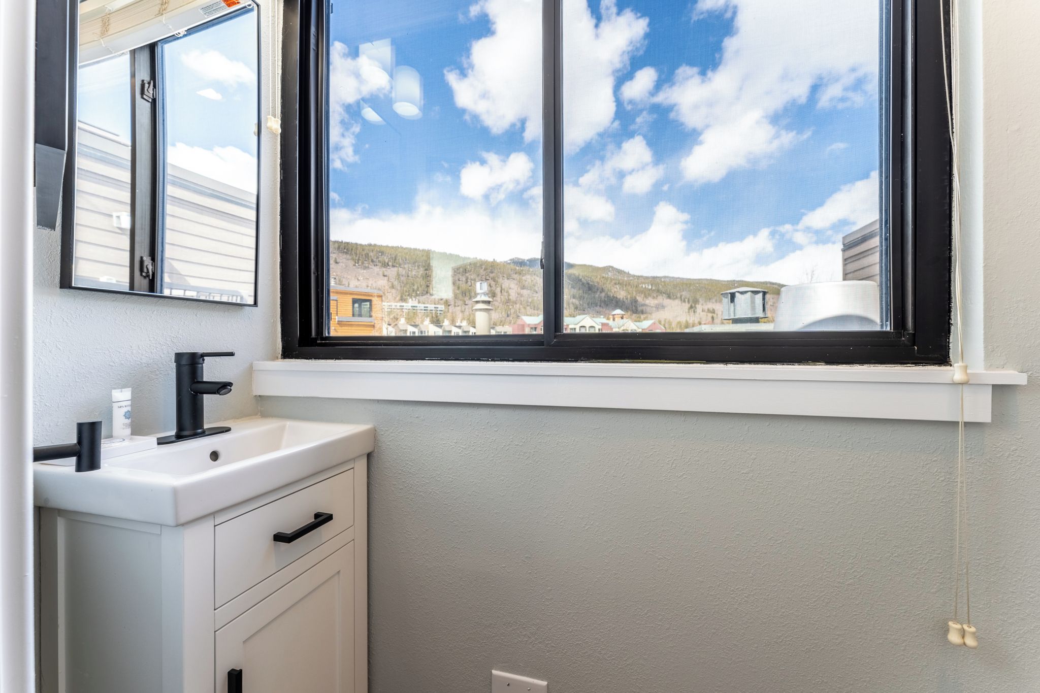 Un baño con lavamanos blanco, grifo negro y ventana con vistas a un pueblo y montañas.