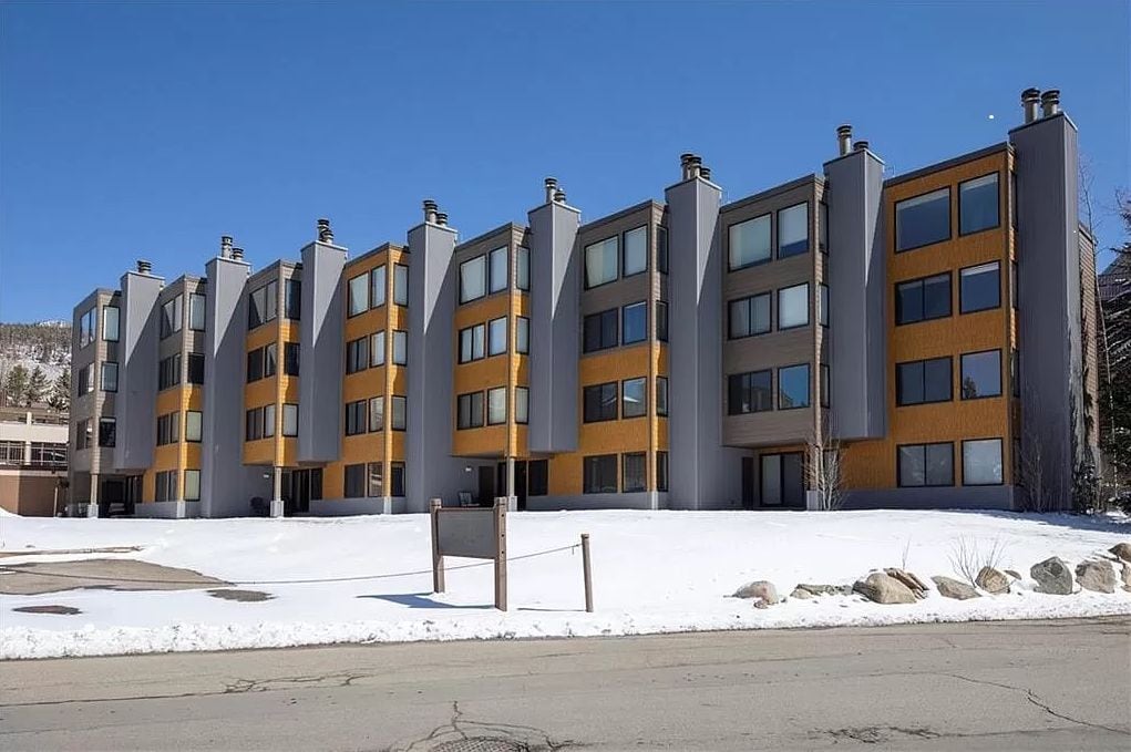 Un hotel de varios pisos con un diseño arquitectónico moderno, que presenta una combinación de fachadas grises y amarillas, ventanas amplias y un suelo cubierto de nieve.