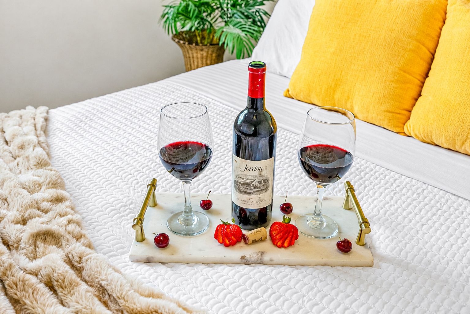 A bed with a white quilted bedspread, a bottle of wine, two glasses of red wine, and a decorative tray with cherries and a strawberry.