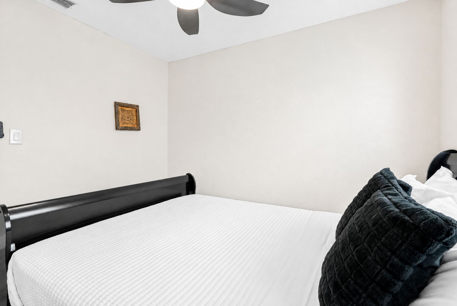 A single bed with a black frame and white bedding, a ceiling fan, and a small framed picture on the wall.