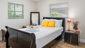 Una cama bien hecha con ropa blanca y almohadas amarillas, una mesita de noche con una lámpara, y una bandeja con copas de vino y una botella.