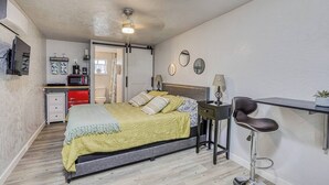 A bedroom with a bed, bedside table, chair, and a small kitchenette area.