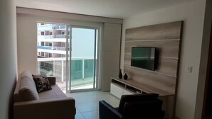 A modern living room with a sofa, a black chair, a TV mounted on a wooden wall, and a sliding glass door leading to a balcony. - 1 bedroom apartment (1 bedroom) Le Bon Vivant (Praia Grande Arraial do Cabo)
