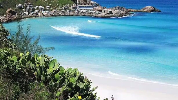 A coastal beach with clear turquoise waters, a sandy shore, and a rocky outcrop with buildings. - 1 bedroom apartment (1 bedroom) Le Bon Vivant (Praia Grande Arraial do Cabo)