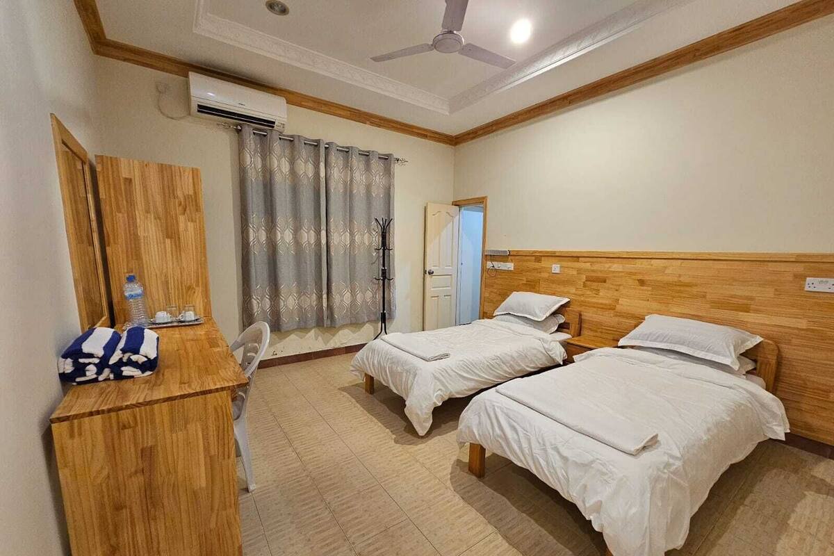 Habitación de hotel con dos camas individuales, un escritorio de madera, una silla, un armario y un ventilador de techo.