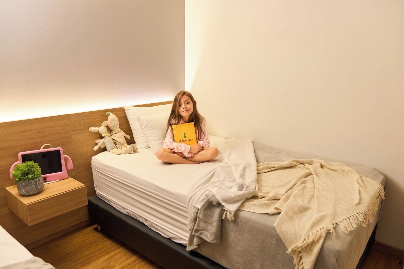 A child sitting on a bed with a book, a tablet, and plush toys on a bedside table.