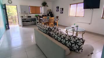A spacious living area with a sofa, dining table, and chairs, a kitchen in the background, and a flat-screen TV mounted on the wall.