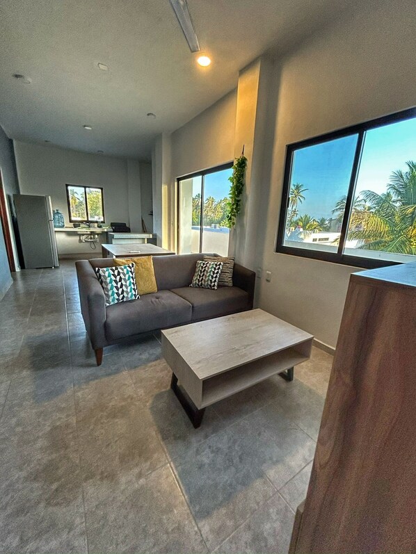 A modern living room with a grey sofa, a wooden coffee table, and a large window with a view of palm trees.