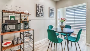 Uma área de refeições com mesa redonda e cadeiras verde-água, uma máquina de café, micro-ondas e uma prateleira com copos e potes.