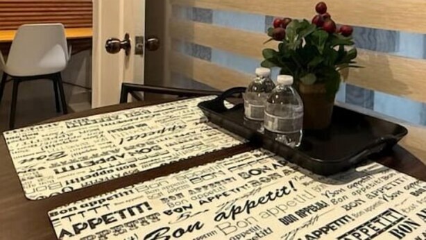 A hotel room with a wooden slatted wall, a dining table with a placemat featuring repeated text, a potted plant, and a tray with water bottles.
