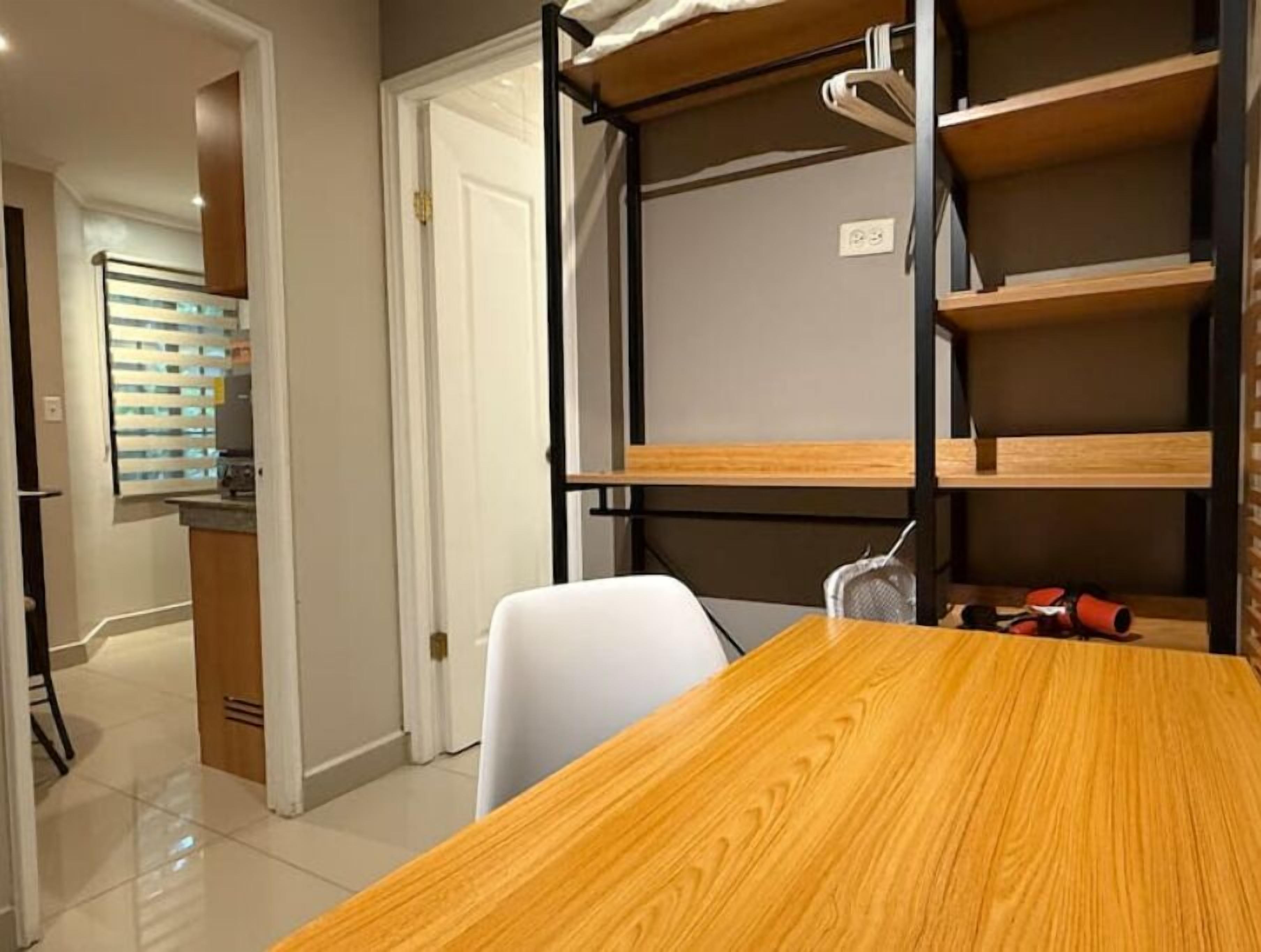 A room with a bunk bed, a wooden table, and a chair. There is a door leading to another room with a window with blinds.