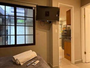 A hotel room with a bed, a TV mounted on the wall, a window with a view of buildings, and a kitchen area visible through an open door. - Lovely Studio Apartment in Zona Bancaria (Panamá)