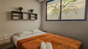 A bedroom with a bed, a nightstand, a shelf with plants, and a window with a palm tree view. - 2-Bedroom Apt in Cinta Costera (Panamá)