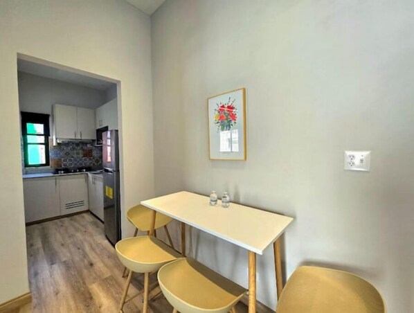 Un pequeño comedor con una mesa y tres sillas, una cocina al fondo y un cuadro en la pared.