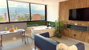 A modern hotel room with a large window, a bathtub, a sofa, a dining area with breakfast, and a TV mounted on a wooden wall. - Rosa Negra Suite w/ Jacuzzi Near Stadium (Medellín)