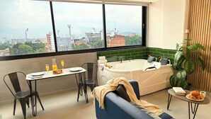 A modern room with a city view, a dining table with breakfast, a sofa, a bathtub, and a potted plant. - Rosa Negra Suite w/ Jacuzzi Near Stadium (Medellín)