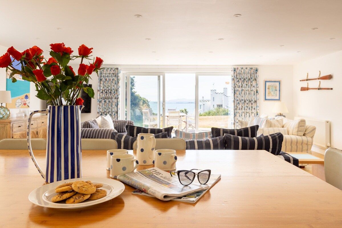 A living room with a wooden table, a vase with red flowers, a plate of cookies, and a newspaper.