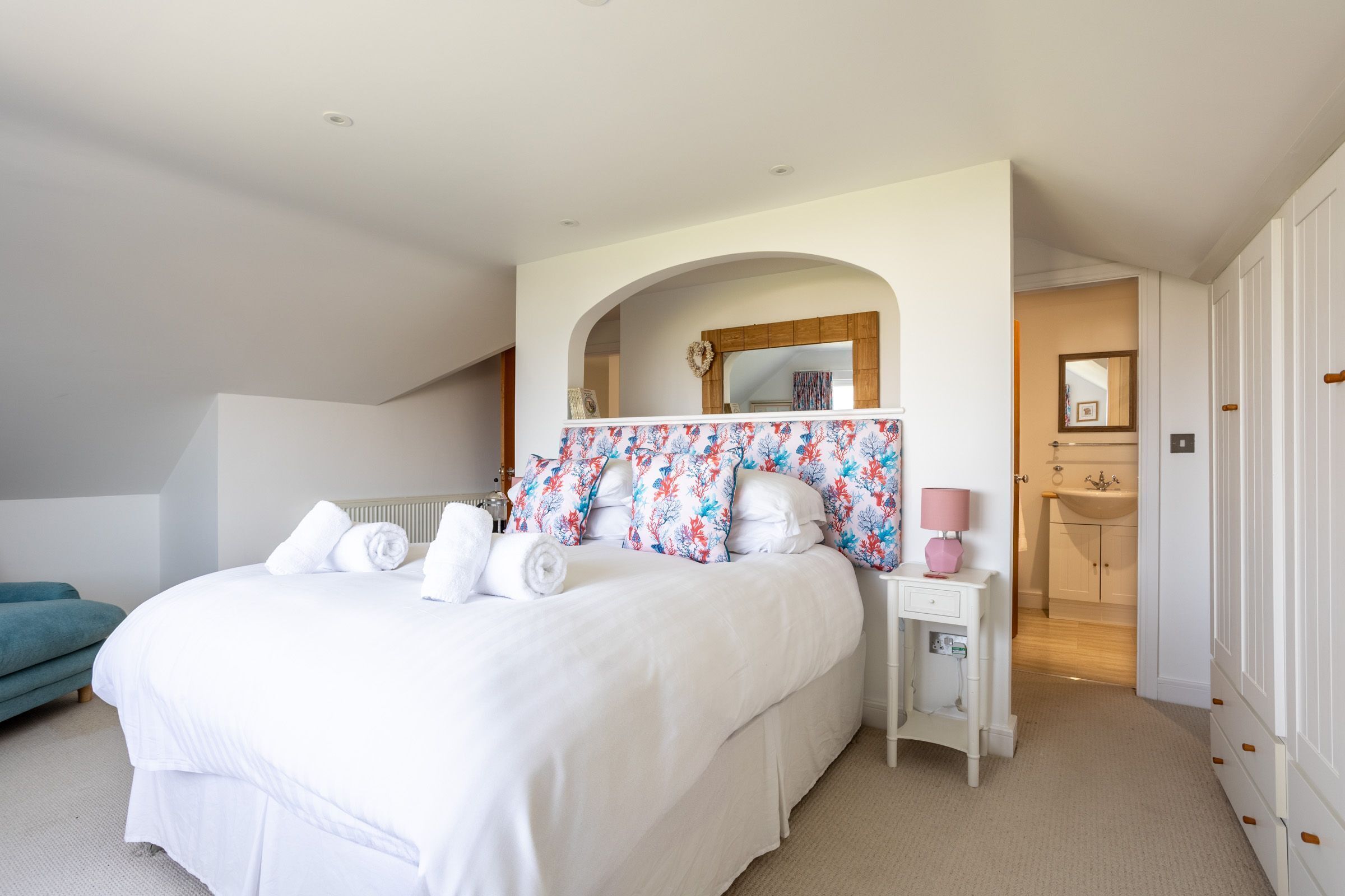 A bedroom with a large bed, a nightstand, a mirror, a bathroom with a sink and mirror, and a teal chair.