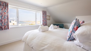 Ein Schlafzimmer mit einem großen Bett, zwei aufgerollten Handtüchern, einem türkisblauen Kopfteil und Blick auf das Meer durch ein Fenster.