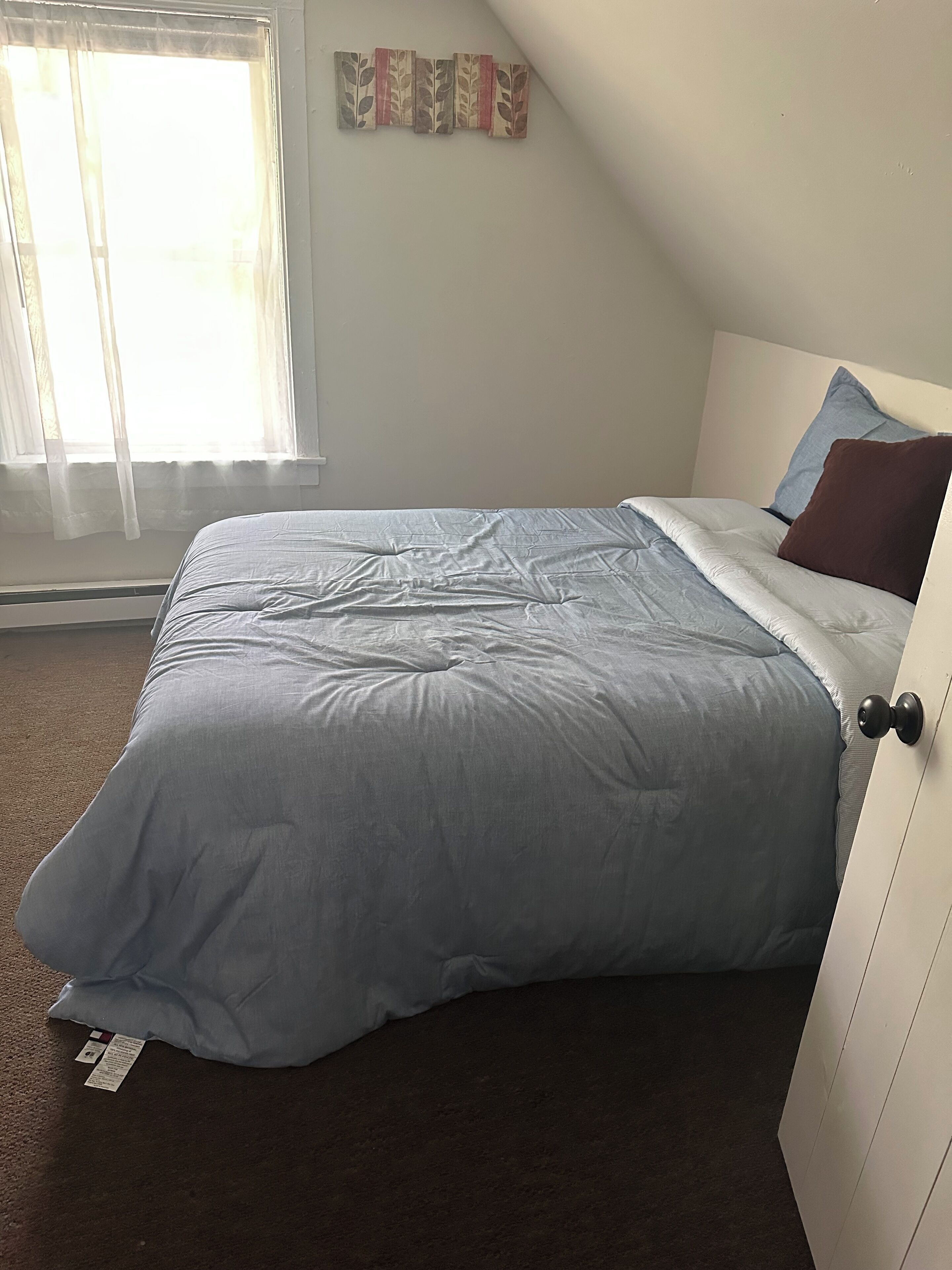 A bedroom with a bed, a window with sheer curtains, a wall-mounted decoration, and a door.