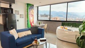 Un salon moderne avec un canapé bleu, une petite table basse et un espace cuisine blanc doté d’une grande fenêtre offrant une vue sur la ville.