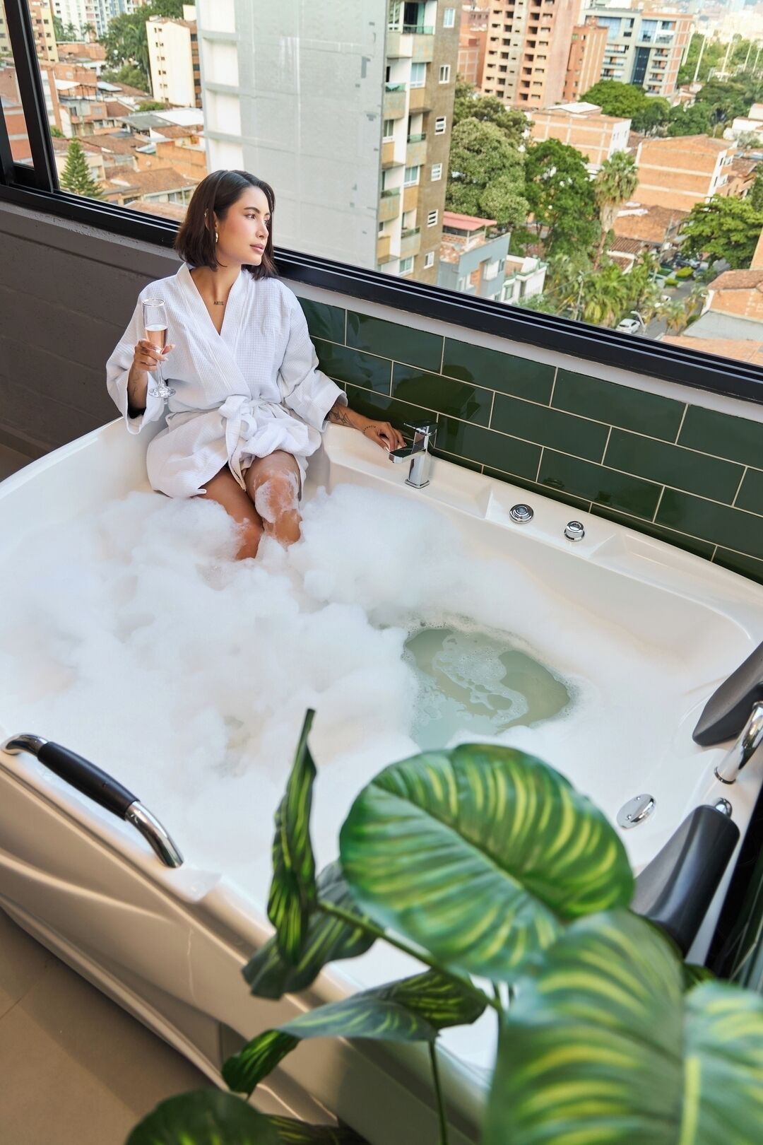 A person sitting in a bathtub filled with bubbles, holding a glass of champagne, with a view of buildings and greenery outside.
