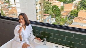 Une personne vêtue d’un peignoir blanc est assise dans une baignoire remplie de mousse, avec une vue sur la ville en arrière-plan.