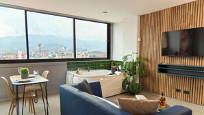 Un salon moderne comprenant un canapé, une table à manger avec des chaises, une grande fenêtre donnant sur la ville, une baignoire et un téléviseur fixé au mur.