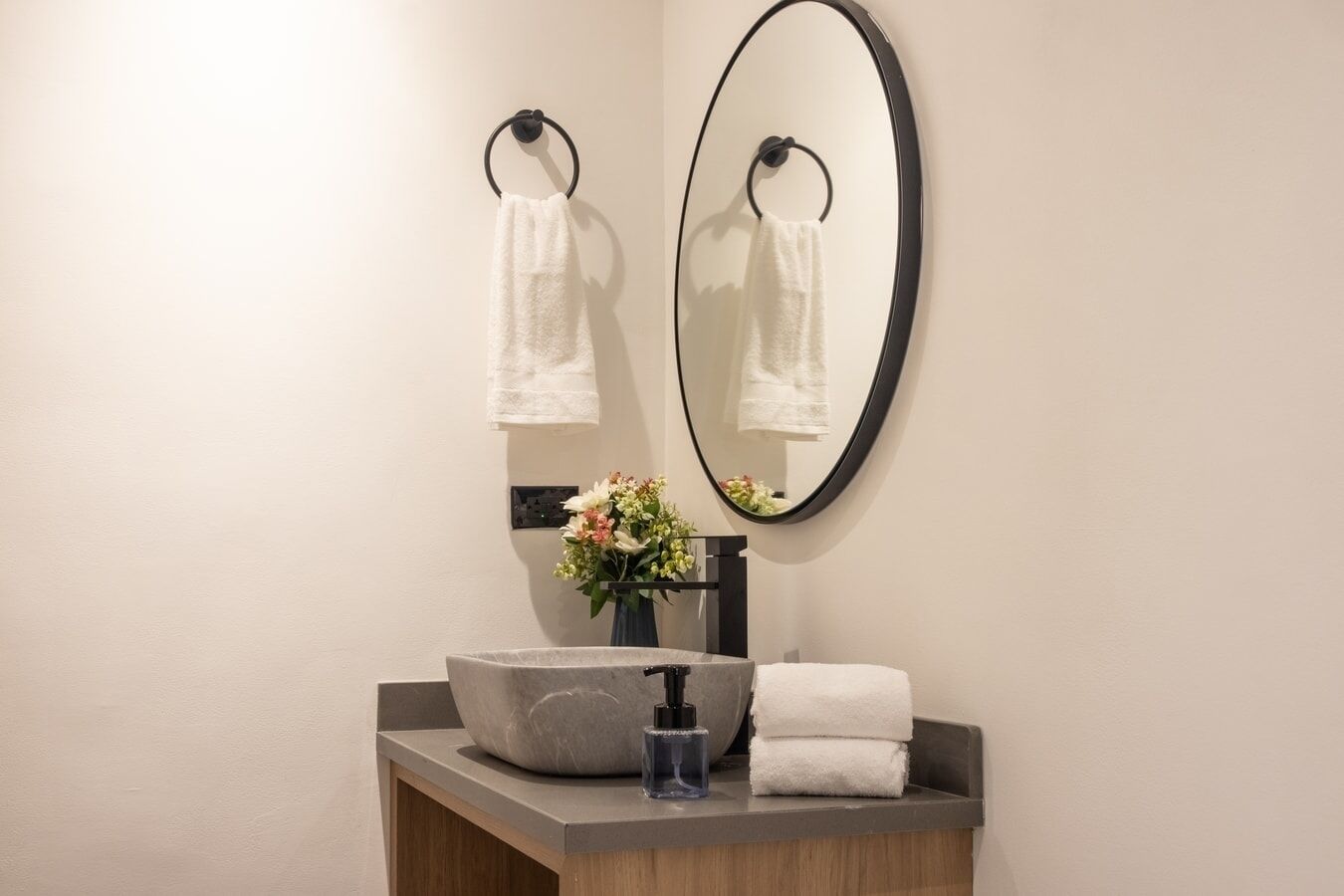 Une salle de bain avec un miroir rond, deux porte-serviettes, un lavabo en pierre, un petit vase avec des fleurs et un distributeur de savon.
