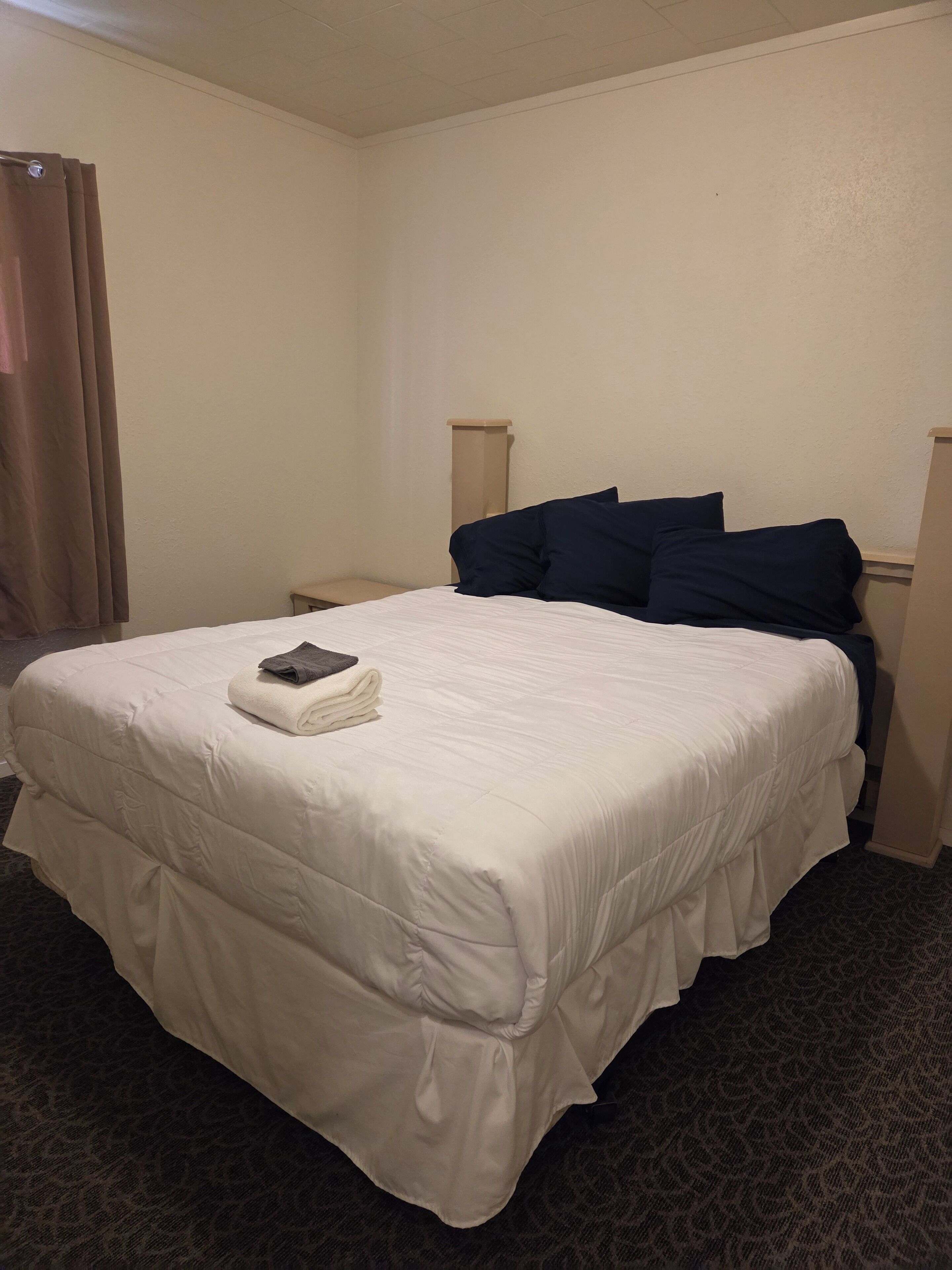 A hotel room with a large bed, a small bedside table, a lamp, and a window with curtains.