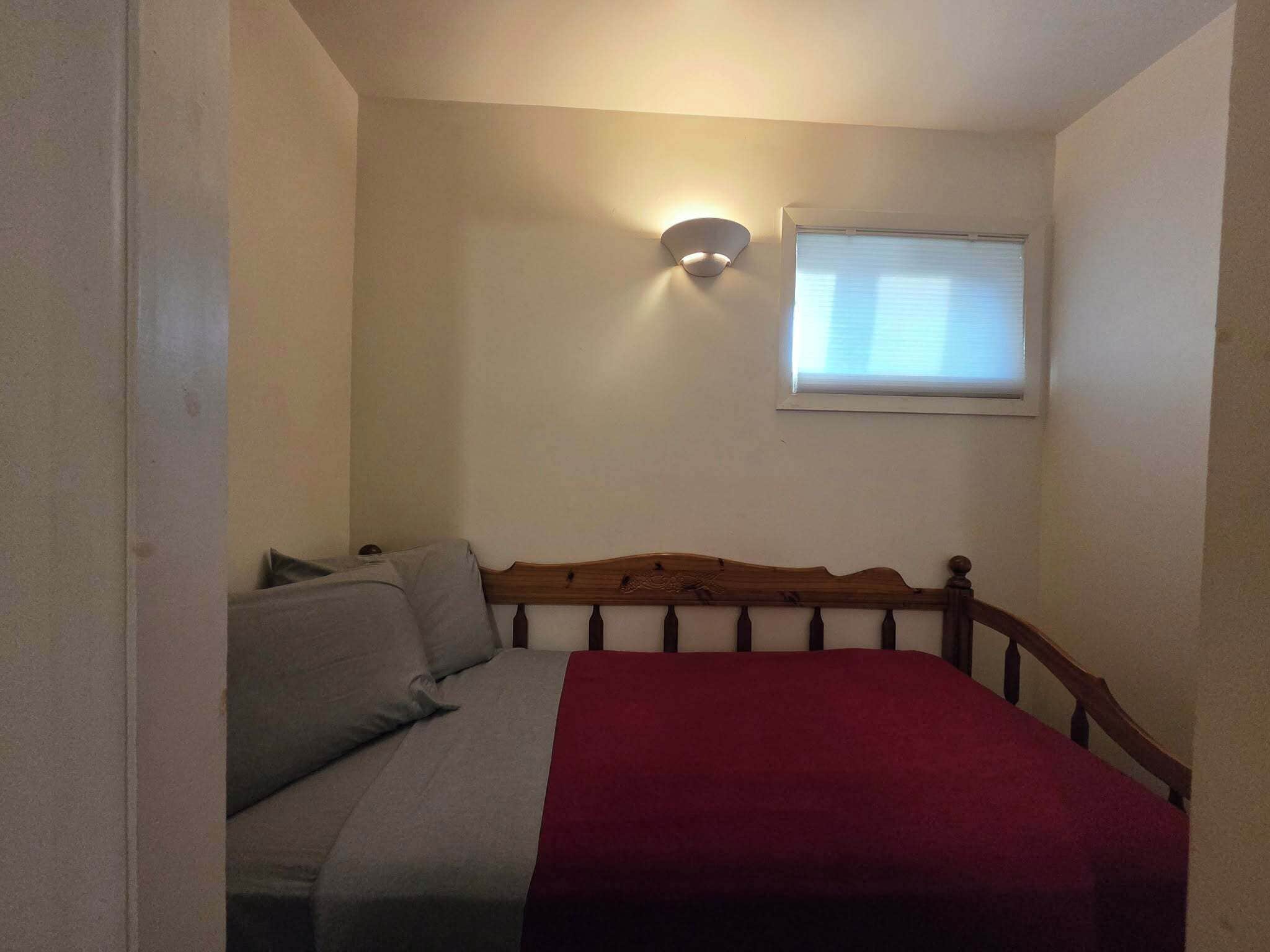 A small, single-bed room with a wooden headboard, a red blanket, and a wall-mounted light fixture.