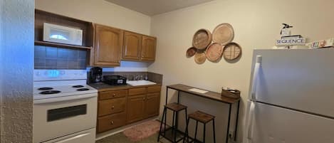 A compact kitchen with wooden cabinets, a white stove, a sink, and a refrigerator. There are two stools and a small table.
